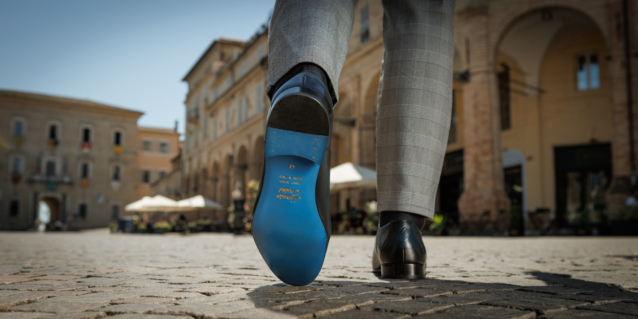 Nunzio Orsini signature blue sole striding across Marche, Italy piazza cobblestones